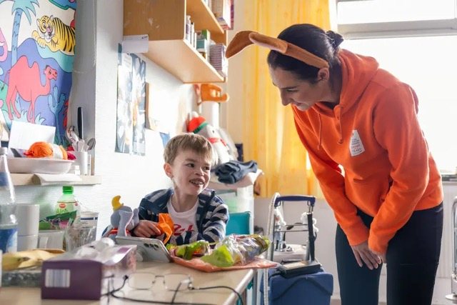 Helferin in orangefarbenem Shirt reicht einem Kind eine Mahlzeit